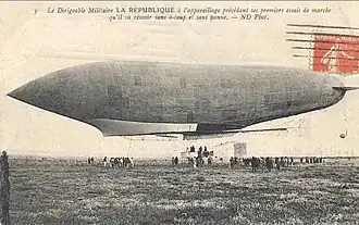 Postcard with orange stamp (French) affixed shows left flank of an airship just above the ground; some people are on the ground below the airship