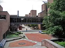 A brick courtyard is flanked by three-story brick buildings with a black glass bridge between them. Trees are visible to the right.