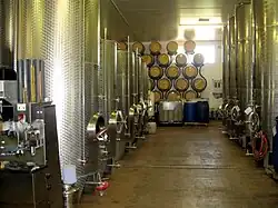 Colour photo showing stainless steel fermentation tanks in the UK. The tanks are located on both sides of an aisle at the bottom of which oak barrels for aging, stacked on a bracket against the wall can be seen. The floor is painted and pitched to a drainage channel in the middle of the winery.