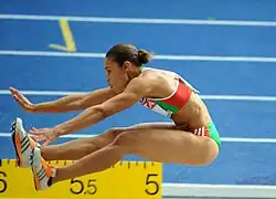 Naide Gomes, in sportwear bearing the green, red, and white of the Portuguese flag, jumps toward the left of the camera, arms and legs stretched forward.