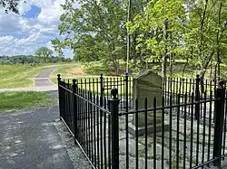 An upright white marble slab in a small paved area surrounded by a black metal fence at the end of an asphalt path through a grass field and around a stand of trees